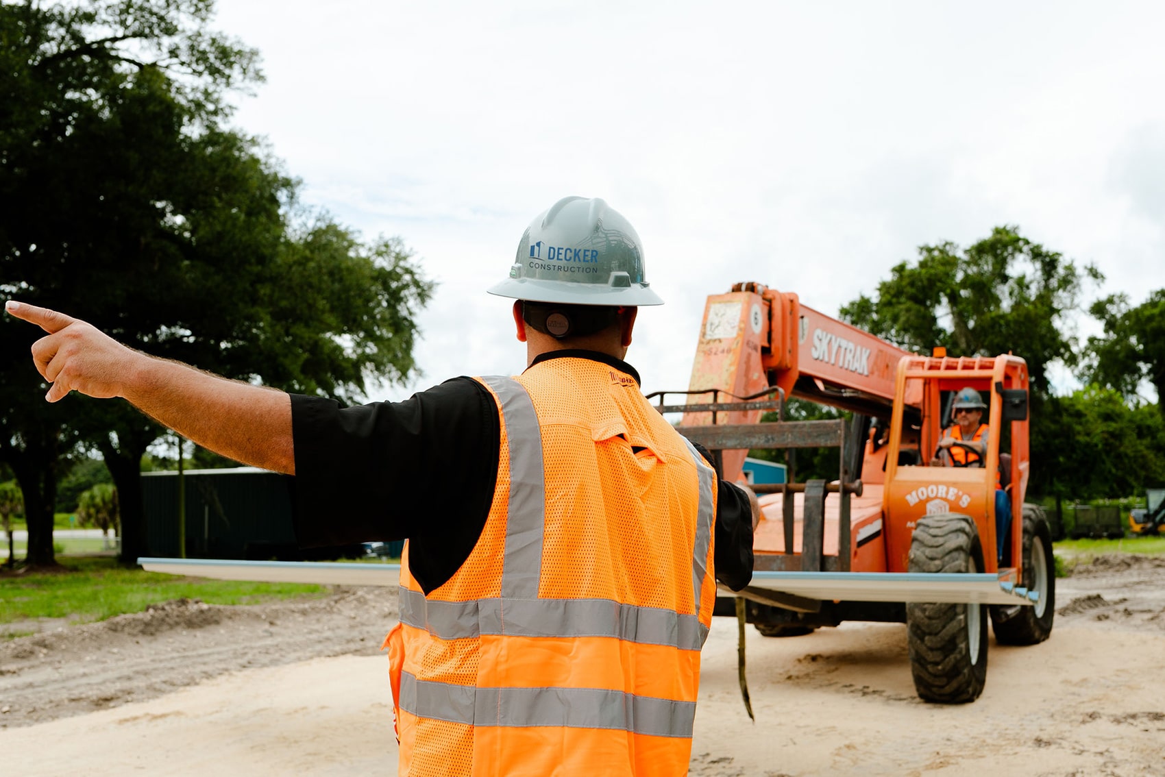 back of a man wearing a safety vest and hard hat motioning an orange tractor where to go